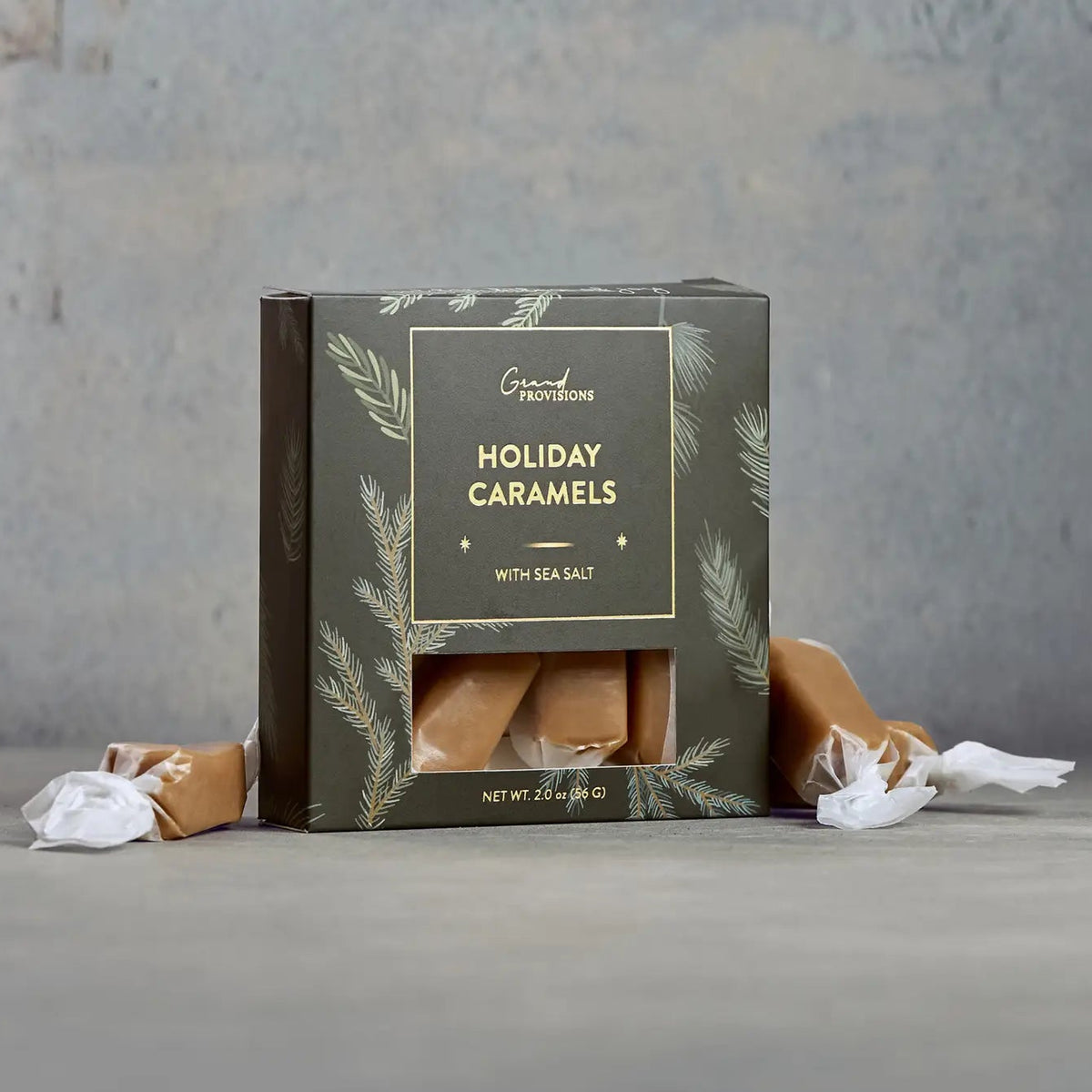 A box of Ground Provisions Holiday Caramels with sea salt, displayed on a gray surface with two unwrapped caramels and white wrapping paper in the foreground.