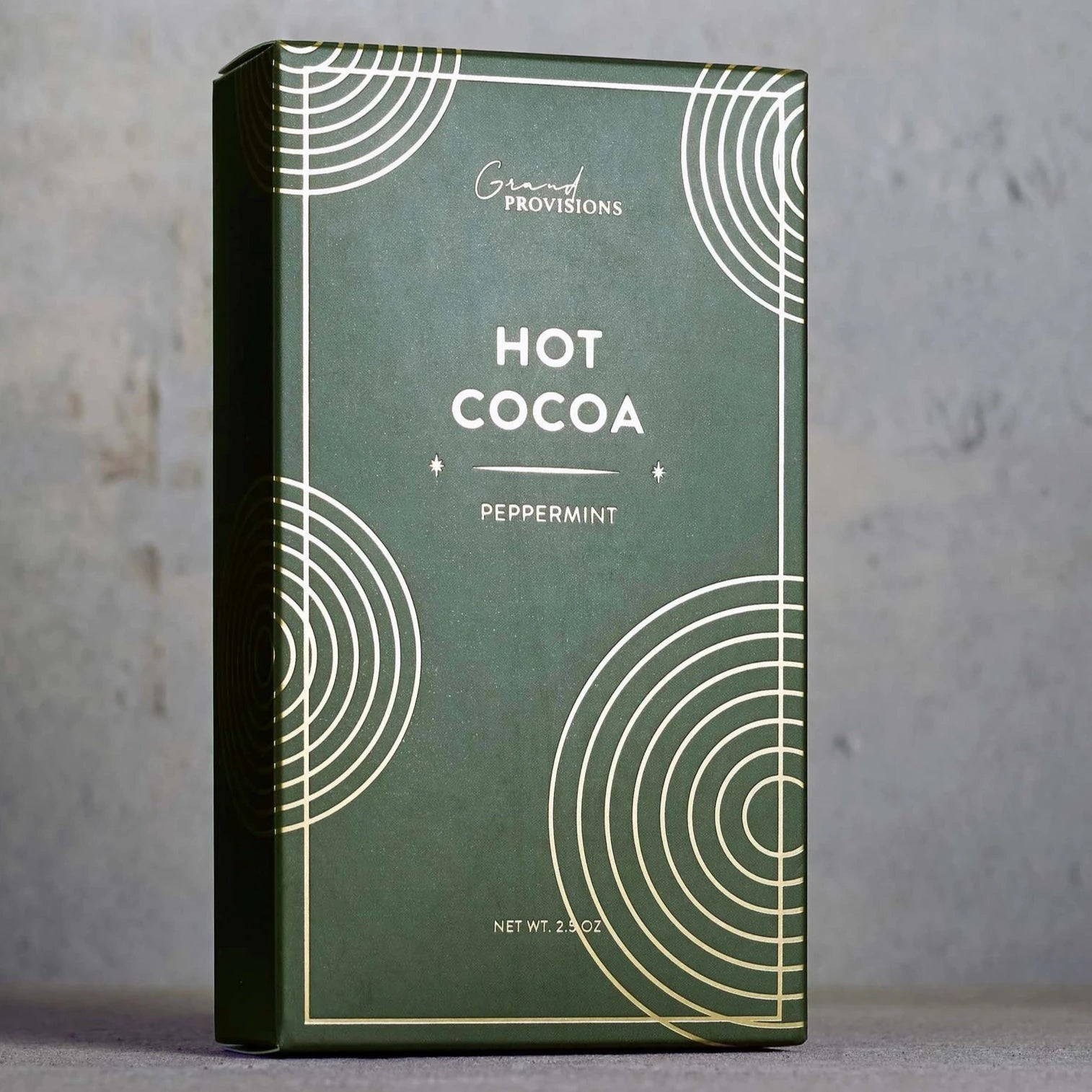 The latest image displays a minimalist and elegant box of "Hot Cocoa" with a peppermint flavor by "Grand Provisions." The packaging utilizes a deep green background with gold geometric circular patterns, creating a sophisticated and refined look. The typography is clean, and the layout feels premium, suggesting this product might be targeted toward a more upscale market or intended as a giftable item.