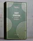 The latest image displays a minimalist and elegant box of "Hot Cocoa" with a peppermint flavor by "Grand Provisions." The packaging utilizes a deep green background with gold geometric circular patterns, creating a sophisticated and refined look. The typography is clean, and the layout feels premium, suggesting this product might be targeted toward a more upscale market or intended as a giftable item.