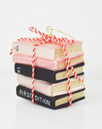 ornament shaped like a stack of book tied together with red and white twine. Book are pink and black with gold glitter showing the pages. 