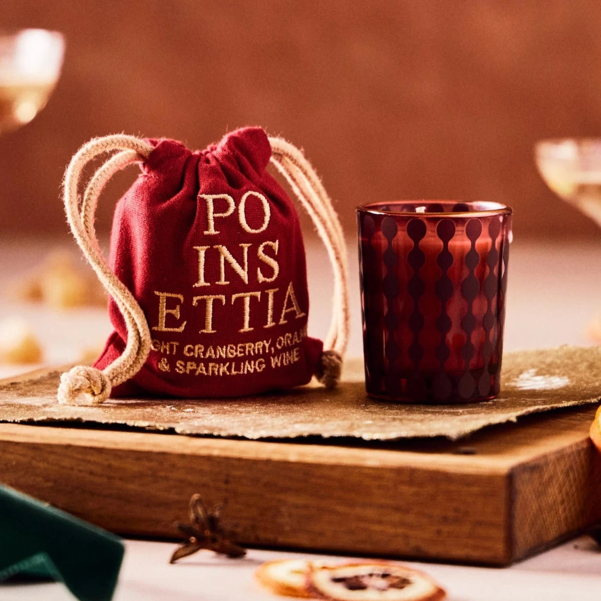 This image shows an elegant setup featuring a decorative glass next to a red fabric pouch with white drawstrings. The pouch has the word "Poinsettia" written in gold letters, along with a description that says "light cranberry, orange & sparkling wine." The background is softly blurred, with cocktail glasses and dried orange slices, creating a warm and festive atmosphere typical of holiday celebrations.