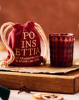 This image shows an elegant setup featuring a decorative glass next to a red fabric pouch with white drawstrings. The pouch has the word "Poinsettia" written in gold letters, along with a description that says "light cranberry, orange & sparkling wine." The background is softly blurred, with cocktail glasses and dried orange slices, creating a warm and festive atmosphere typical of holiday celebrations.