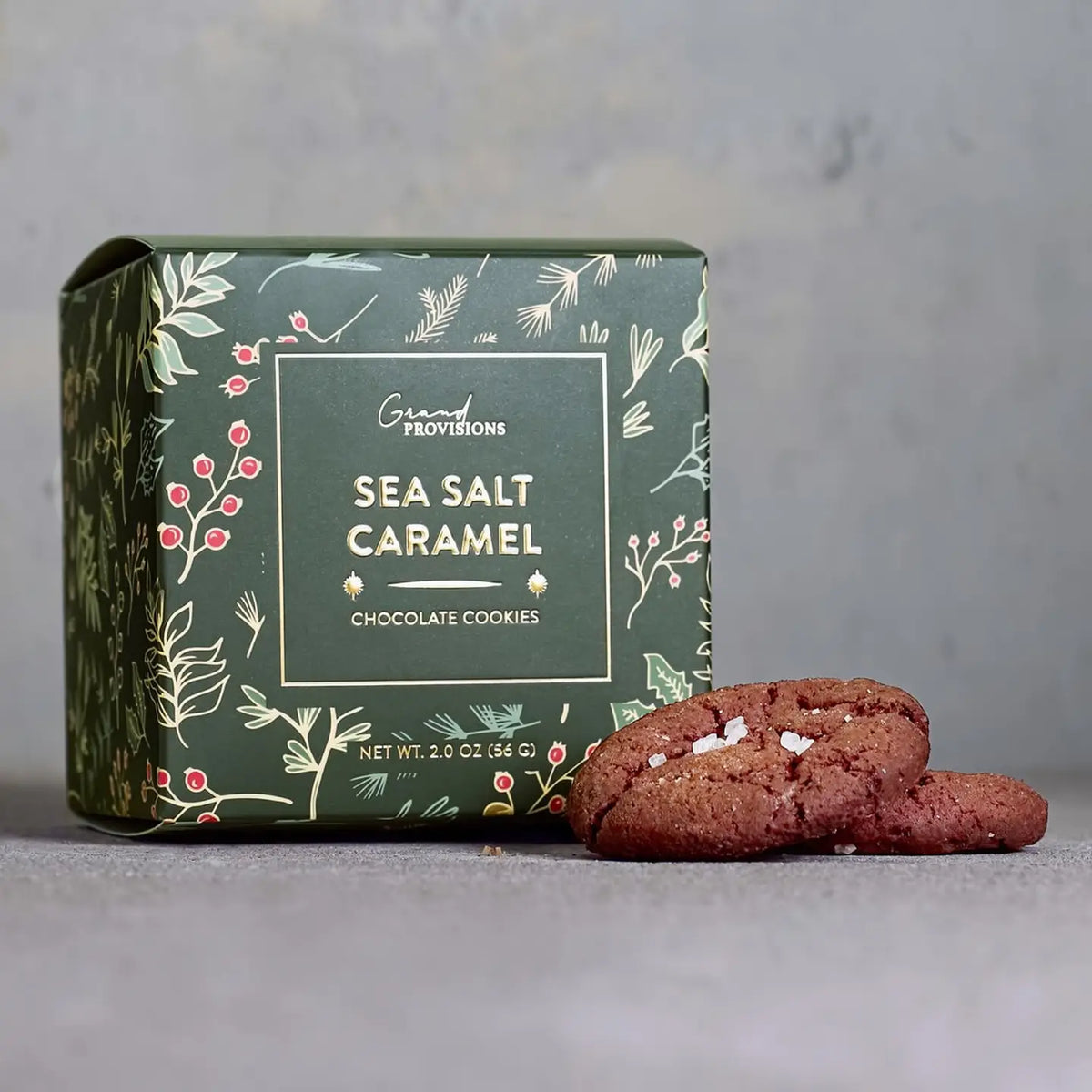 A box of Grand Provisions Sea Salt Caramel Chocolate Cookies sits on a gray surface with three cookies placed in front. The box is dark green with botanical illustrations and white text, and the cookies are brown with visible sea salt crystals.