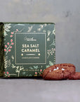 A box of Grand Provisions Sea Salt Caramel Chocolate Cookies sits on a gray surface with three cookies placed in front. The box is dark green with botanical illustrations and white text, and the cookies are brown with visible sea salt crystals.