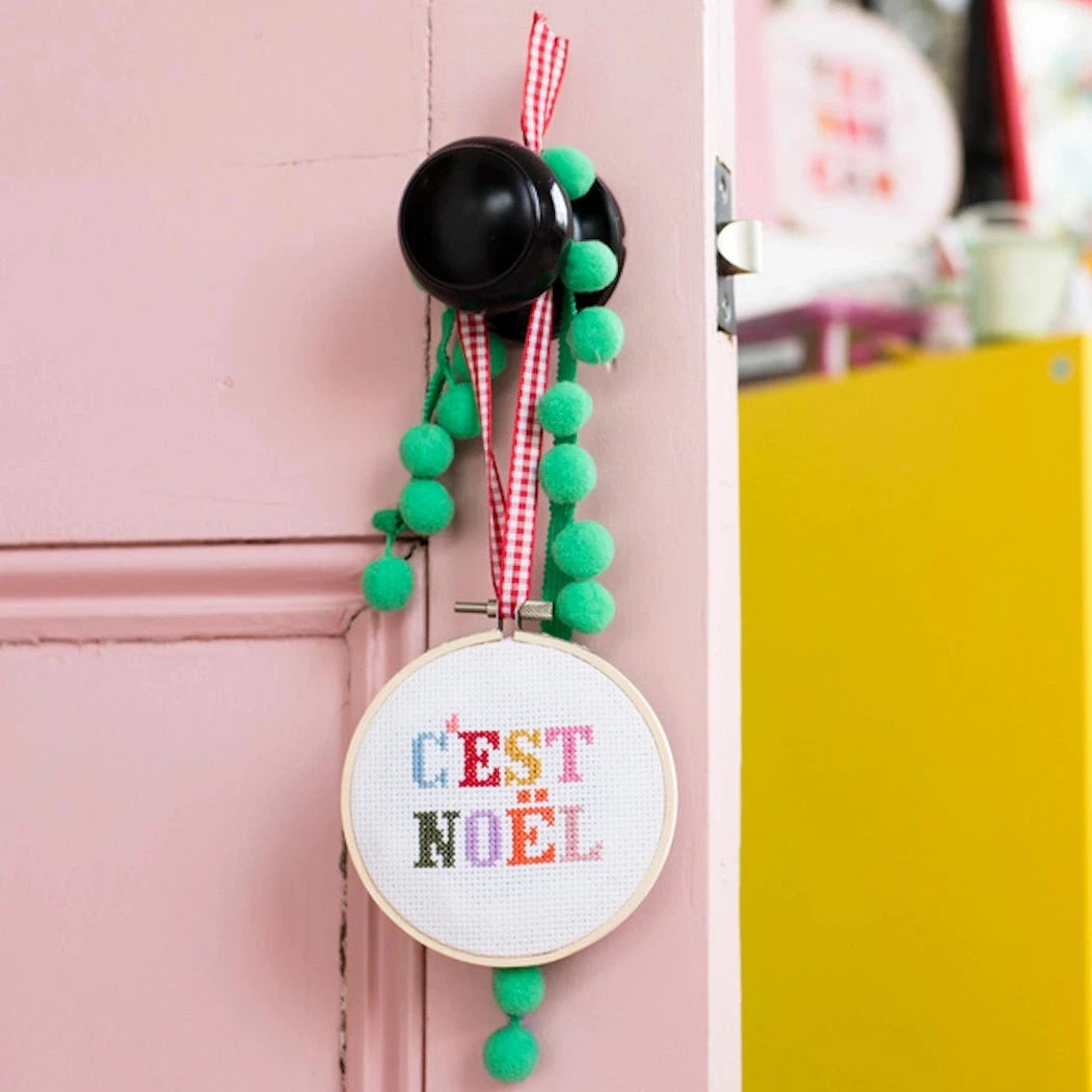 This image shows the same "C'EST NOËL" cross-stitch hoop, now styled as a festive door ornament. It is hung from a black doorknob using a red-and-white gingham ribbon, with green pom-pom trim adding a playful, holiday touch. The setting includes a pastel pink door, giving it a cheerful, whimsical vibe, and the background shows a bright and colorful space with yellow storage units and decorative items, suggesting a cozy, festive environment.