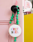 This image shows the same "C'EST NOËL" cross-stitch hoop, now styled as a festive door ornament. It is hung from a black doorknob using a red-and-white gingham ribbon, with green pom-pom trim adding a playful, holiday touch. The setting includes a pastel pink door, giving it a cheerful, whimsical vibe, and the background shows a bright and colorful space with yellow storage units and decorative items, suggesting a cozy, festive environment.