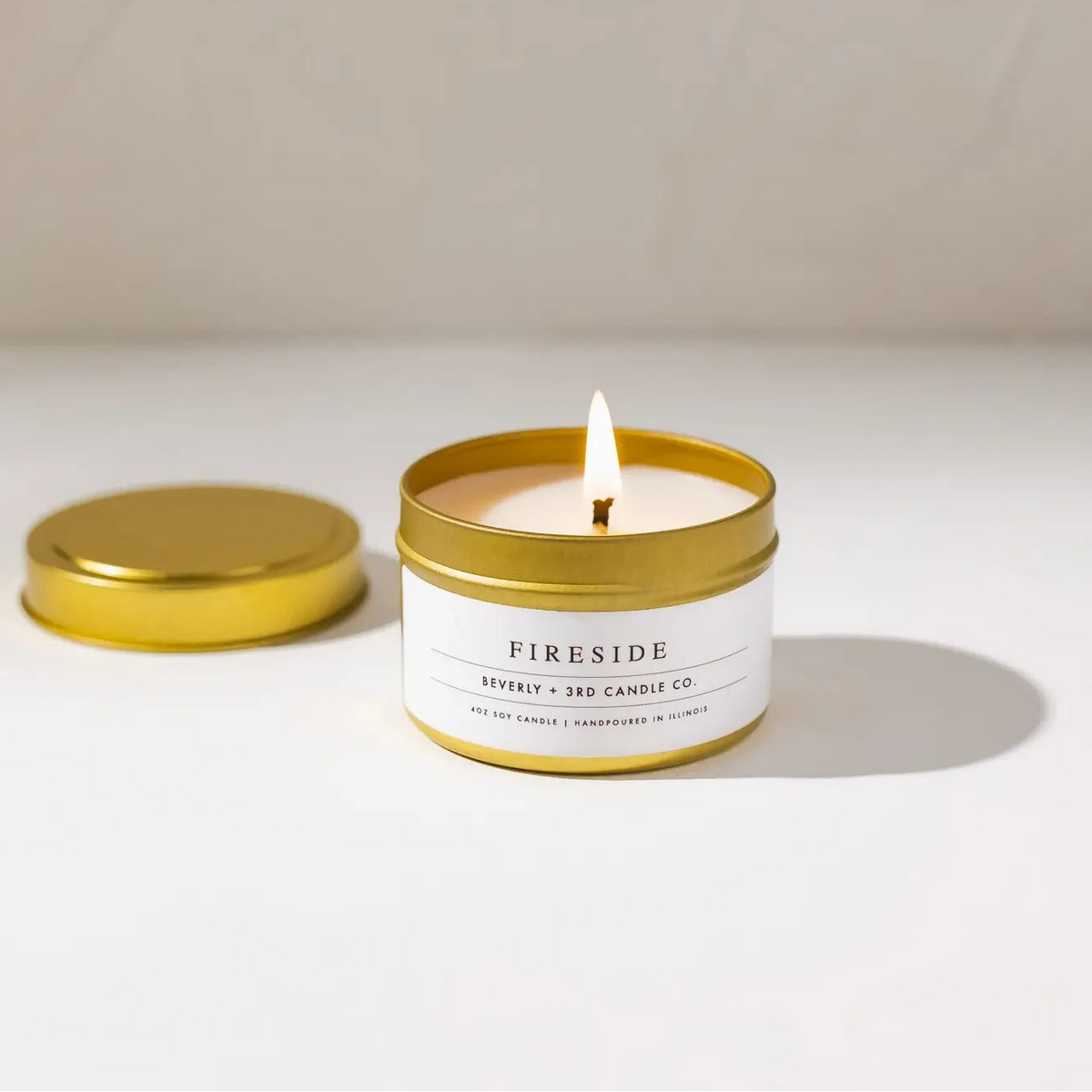 A lit candle in a gold metal tin with its lid placed beside it on a clean white surface. The label reads 'FIRESIDE' and 'BEVERLY + 3RD CANDLE CO.