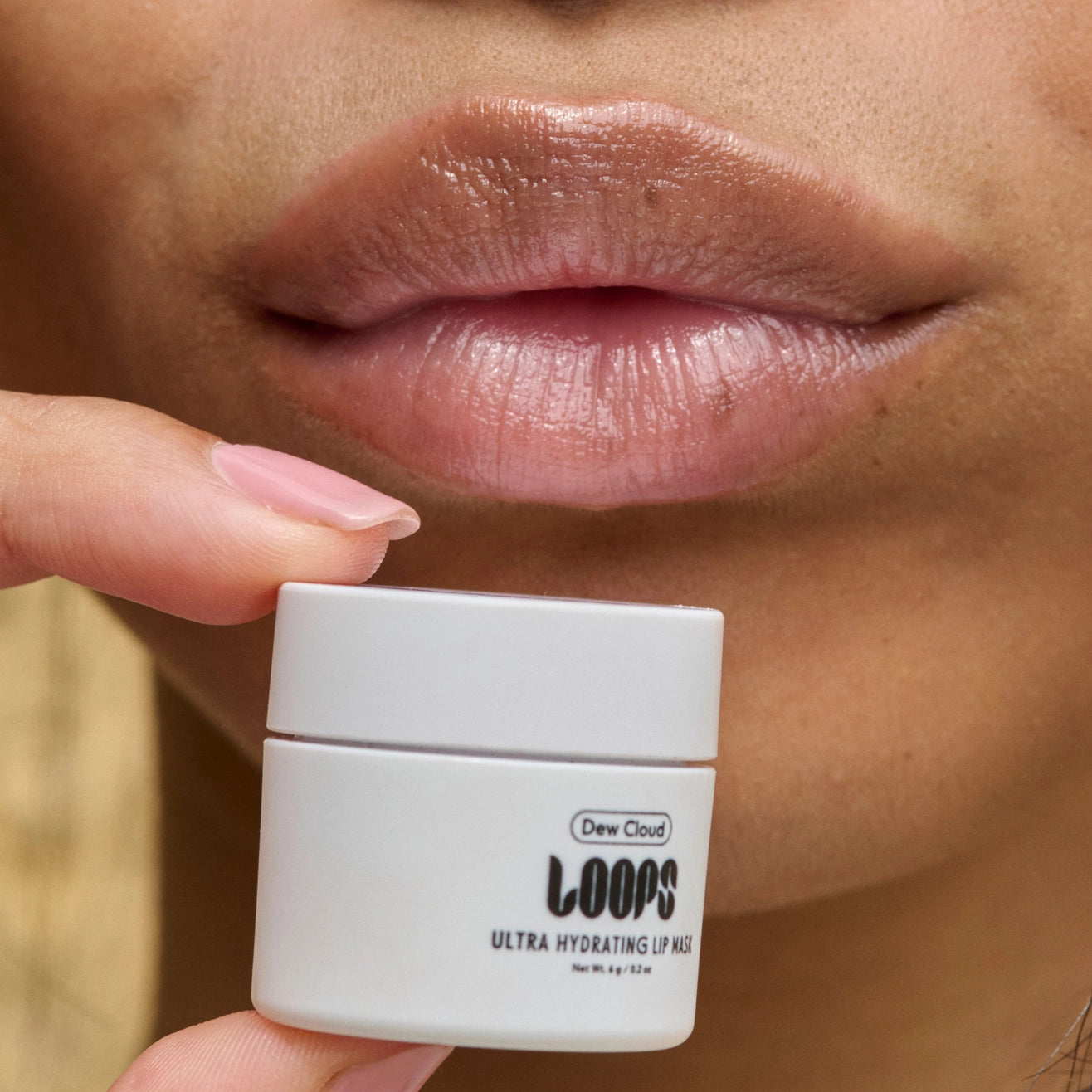 Person holding a white jar of 'LOOPS Ultra Hydrating Lip Wax' near their lips.