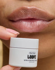 Person holding a white jar of 'LOOPS Ultra Hydrating Lip Wax' near their lips.