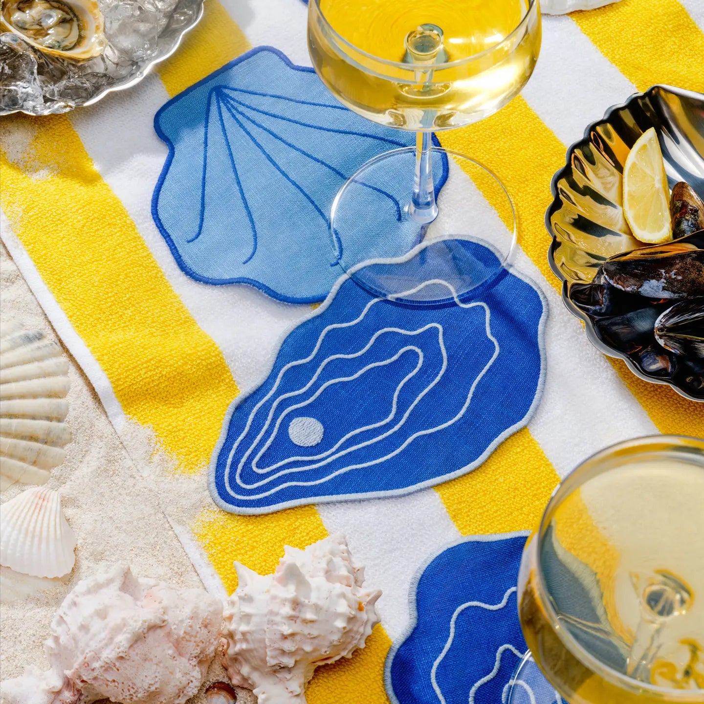 Blue oyster-shaped coasters on a yellow and white striped tablecloth with wine glasses and shells.