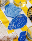 Blue oyster-shaped coasters on a yellow and white striped tablecloth with wine glasses and shells.