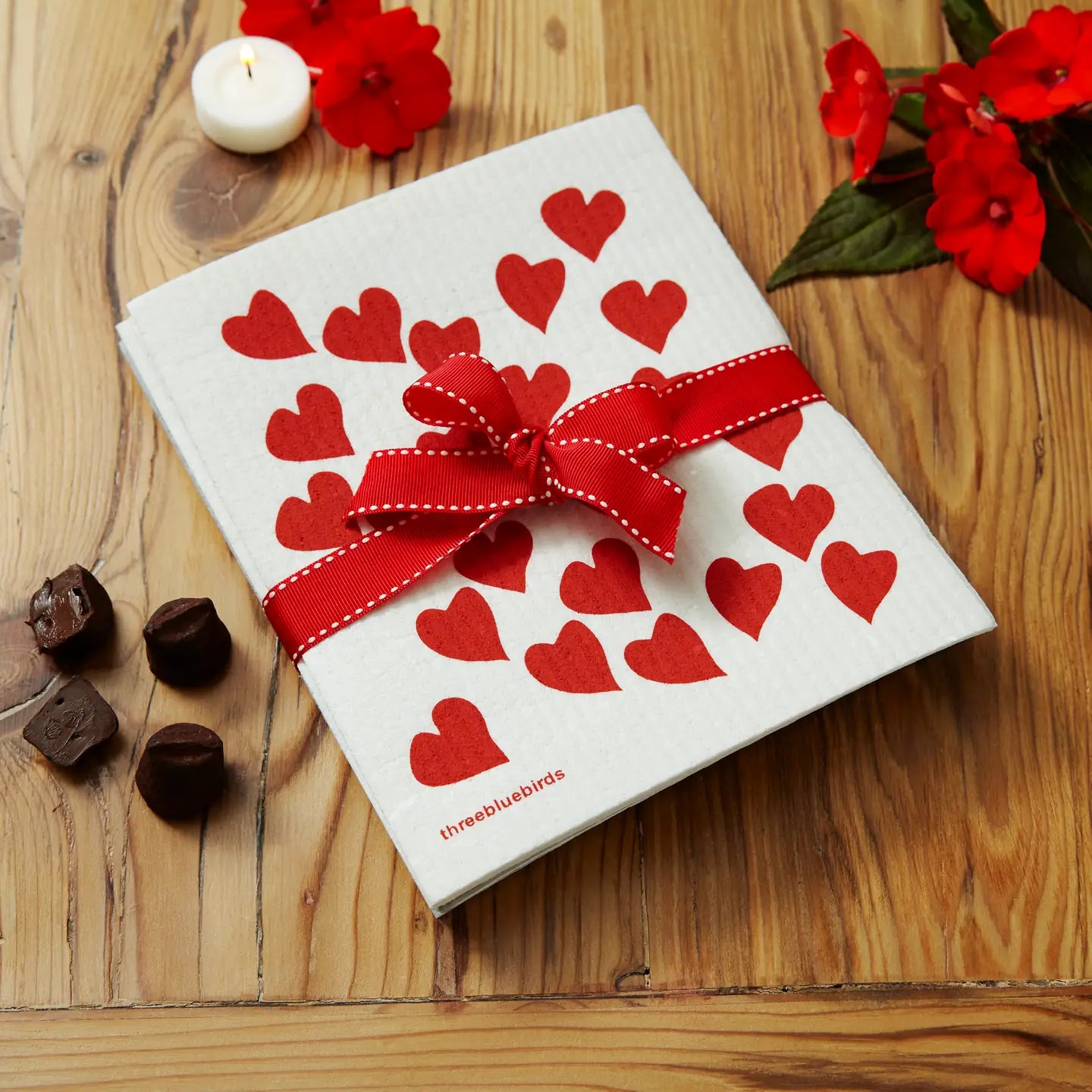 dishcloth with red hearts on it tied with a red bow on a wooden countertop next to red flowers, candles, and chocolates