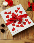 dishcloth with red hearts on it tied with a red bow on a wooden countertop next to red flowers, candles, and chocolates