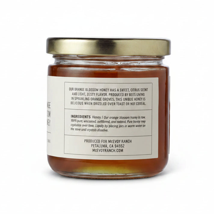 A glass jar of Mevyo Ranch Orange Blossom Honey with a gold metal lid and a white label showing the back text, set against a plain white background.