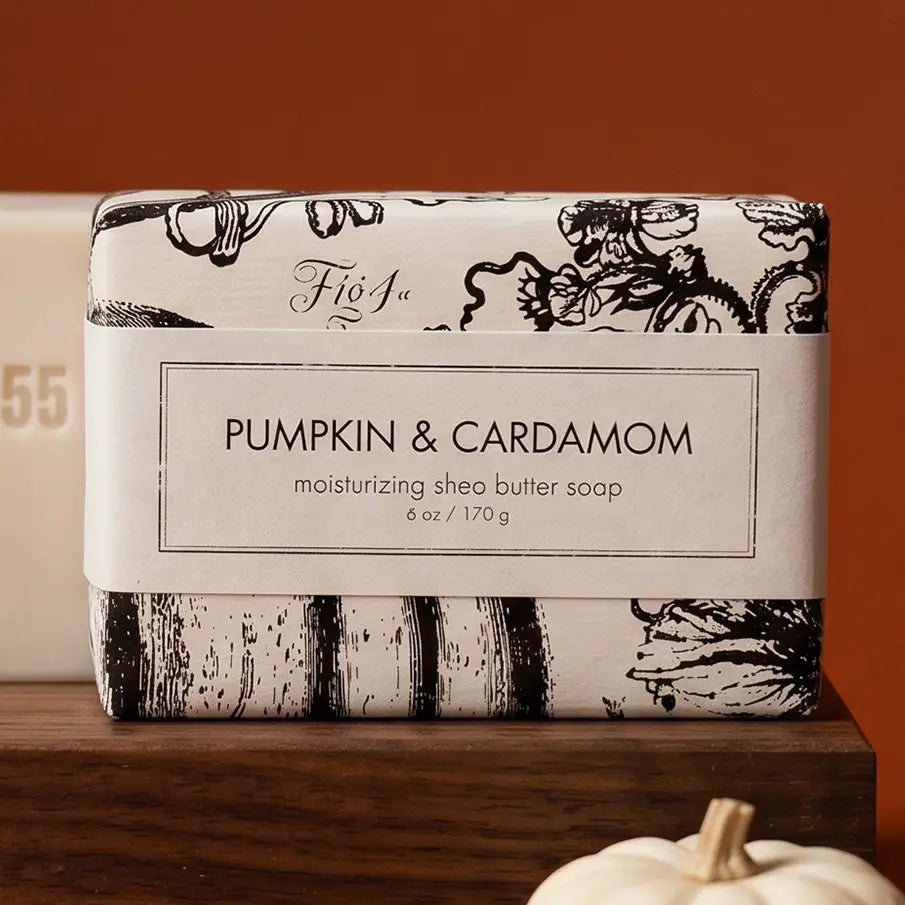 A wrapped bar of Fíña Pumpkin & Cardamom moisturizing shea butter soap sits on a wooden surface against a warm orange-brown background. The soap is wrapped in a white paper band with black text and black botanical/pumpkin illustrations on a cream background.