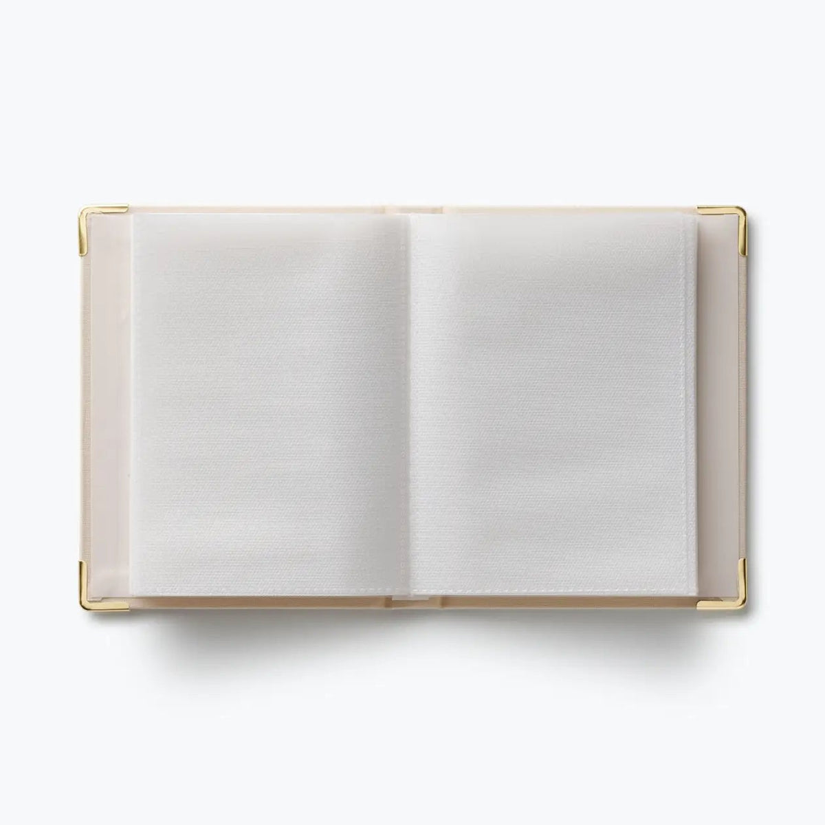 An open hardcover notebook with blank white pages and a light beige cover, framed by gold-toned metal corner protectors, shot from a top-down perspective on a white background.