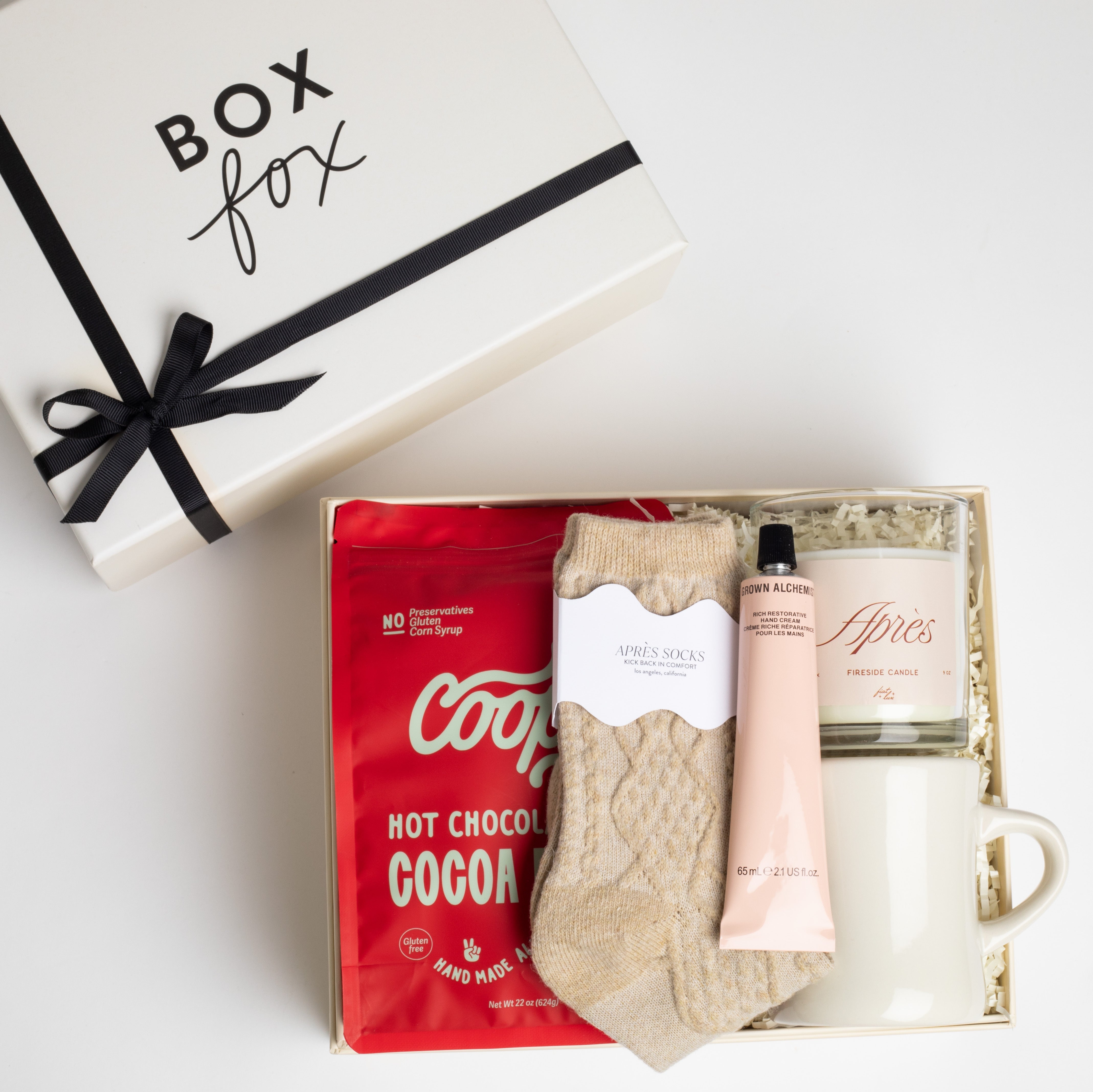 CREME BOXFOX BOX DISPLAYED OPEN WITH: Cocoa Felice Hot Chocolate, Beige Après Ski Socks, Chocolate Covered Cookie Dough Bites, Après Candle, Rich Restorative Hand Cream, and Finders Keepers Coffee Mug