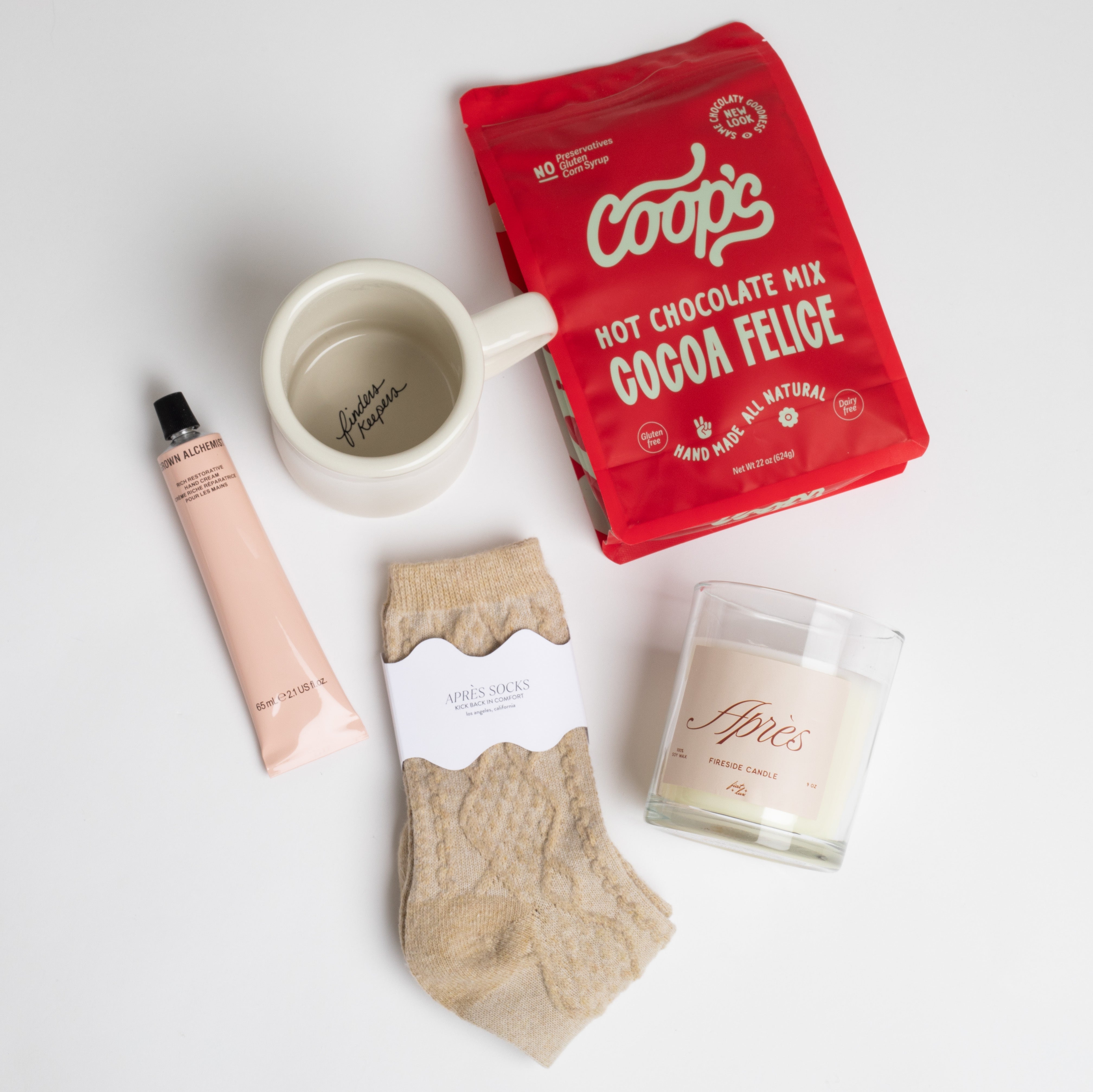 FIRESIDE CURATED BOX ITEMS DISPLAYED: Cocoa Felice Hot Chocolate, Beige Après Ski Socks, Chocolate Covered Cookie Dough Bites, Après Candle, Rich Restorative Hand Cream, and Finders Keepers Coffee Mug