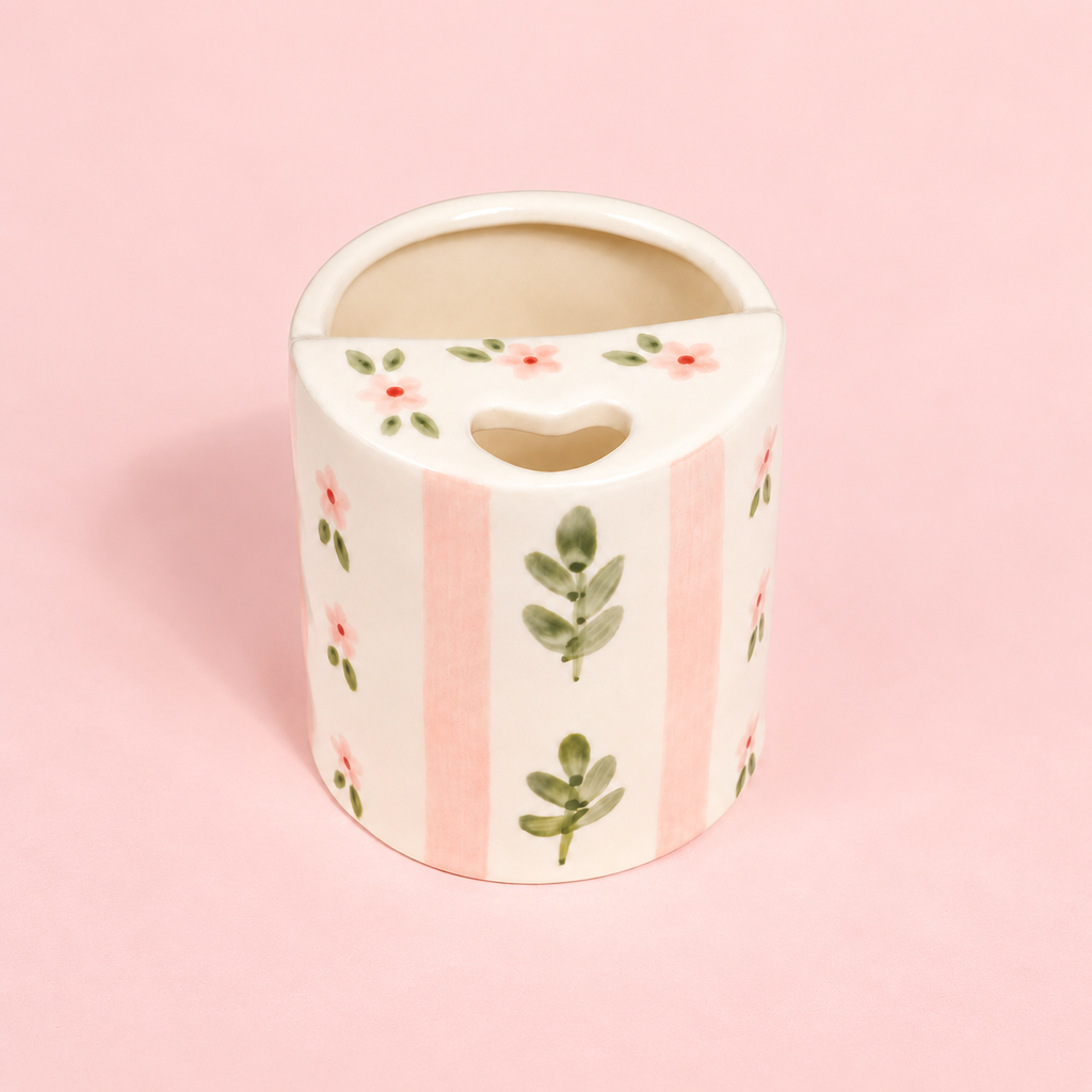 A ceramic tooth holder with a heart-shaped cutout in the center, decorated with pink stripes, green leaves, and small pink flowers on a light pink background.