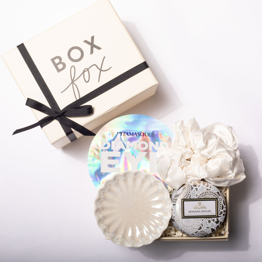 A luxury gift box set from Box Fox, featuring a cream-colored gift box with a black ribbon, and an open tray containing a scalloped cream ceramic dish, a white silk scrunchie, a round silver and black patterned soap or candle, and a holographic round sticker.