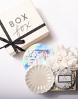 A luxury gift box set from Box Fox, featuring a cream-colored gift box with a black ribbon, and an open tray containing a scalloped cream ceramic dish, a white silk scrunchie, a round silver and black patterned soap or candle, and a holographic round sticker.