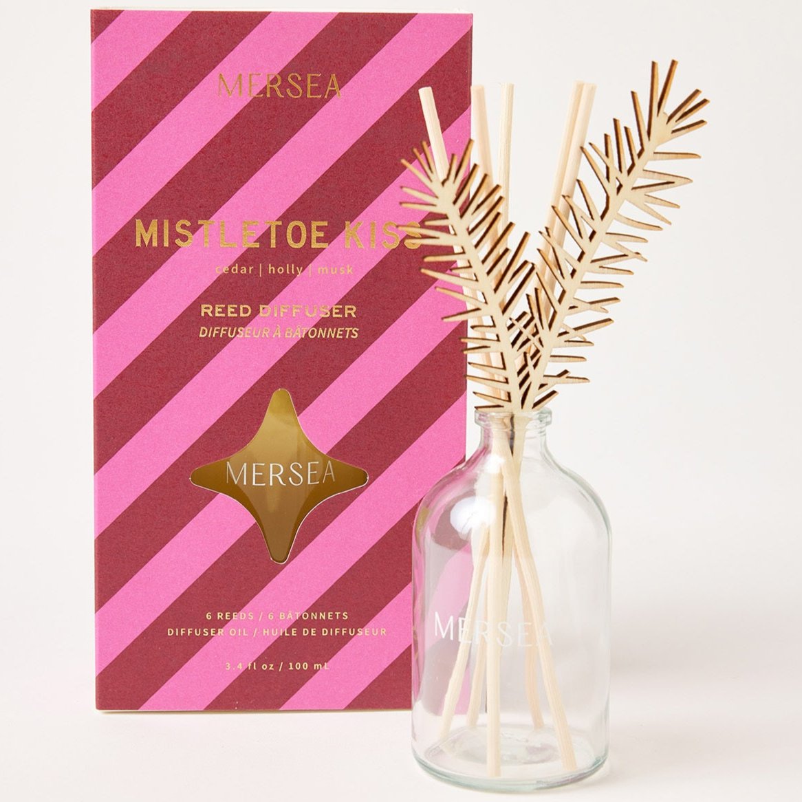 A glass reed diffuser bottle labeled "MERSEA" is placed in front of a red and pink diagonal striped box. The box reads "Mistletoe Kiss" and features notes of cedar, holly, and musk. The reed diffuser comes with six wooden reeds resembling pine needles.