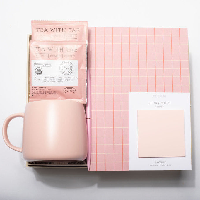 A pink gift set featuring a matte blush pink ceramic mug, a pink tea packet labeled 'TEA WITH TAI', a pink-and-white checkered notebook, and a white card labeled 'STICKY NOTES' on a white background.