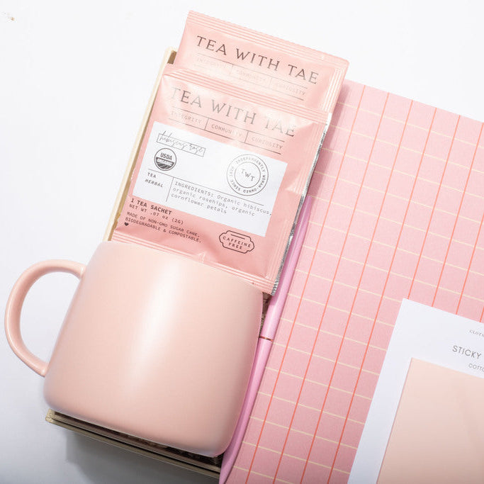 A pink ceramic mug sits next to a pink notebook with a grid pattern, a pink tea packet labeled 'TEA WITH TAE', and a sheet of sticky notes on a white background.
