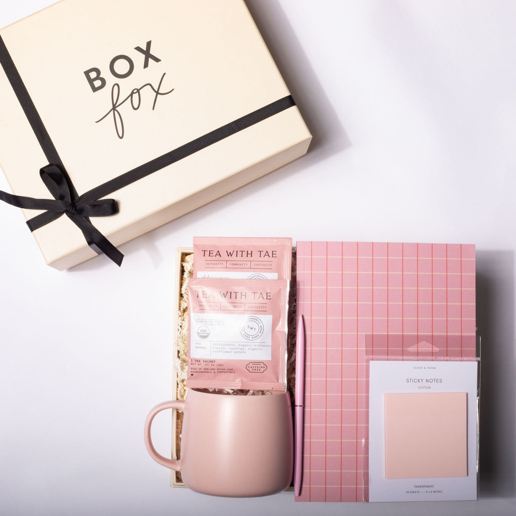 A styled gift set on a white background featuring a cream-colored 'Box Fox' gift box with a black ribbon, a pink ceramic mug, a pink tea packet labeled 'Tea with Tae', a pink grid notebook, and a pink sticky note pad.
