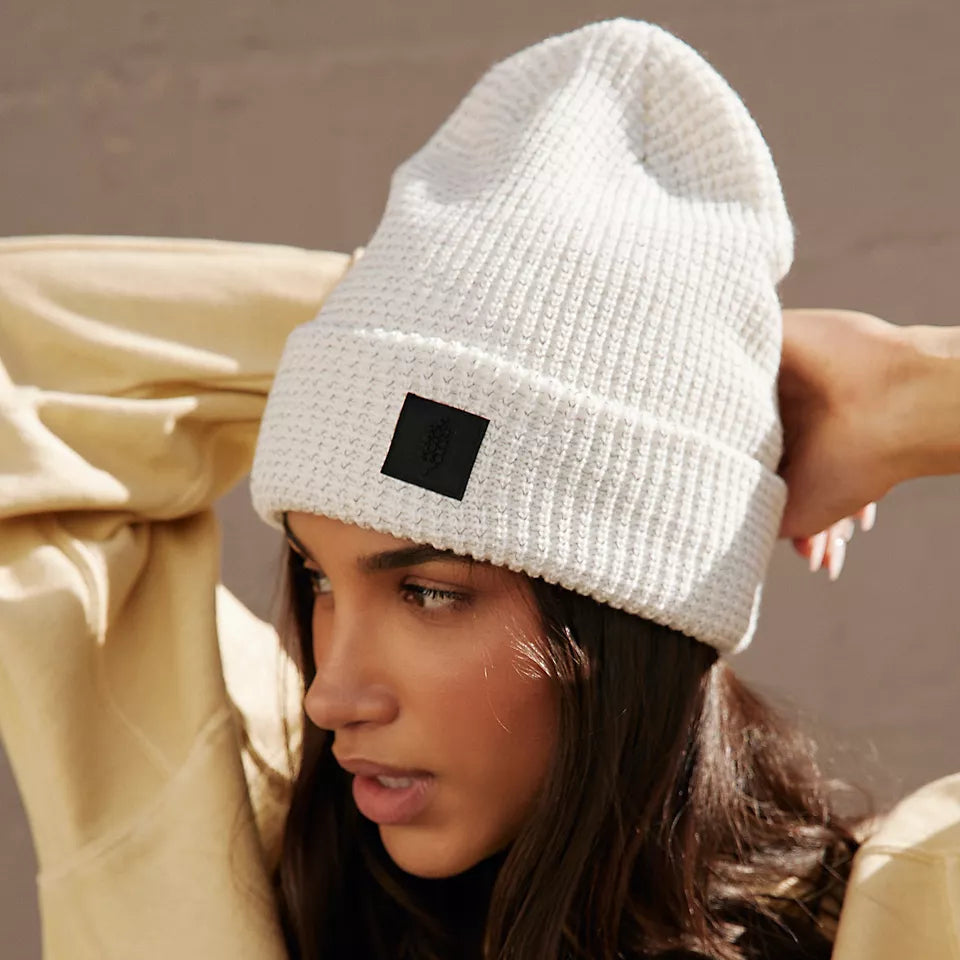 Person wearing a white beanie with a black patch, sitting against a neutral background