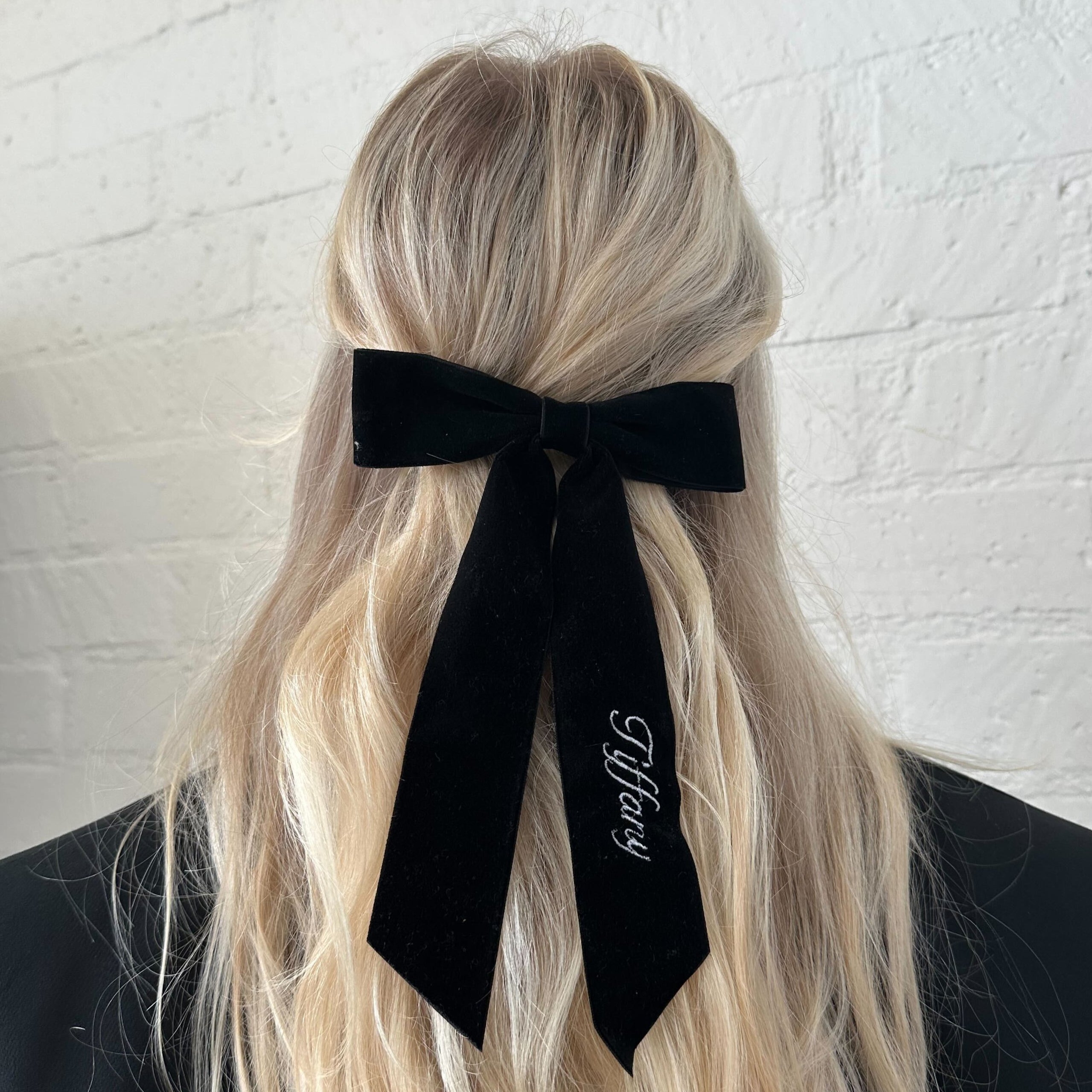 Person with long blonde hair wearing a black bow with 'Tiffany' branding against a white brick wall.
