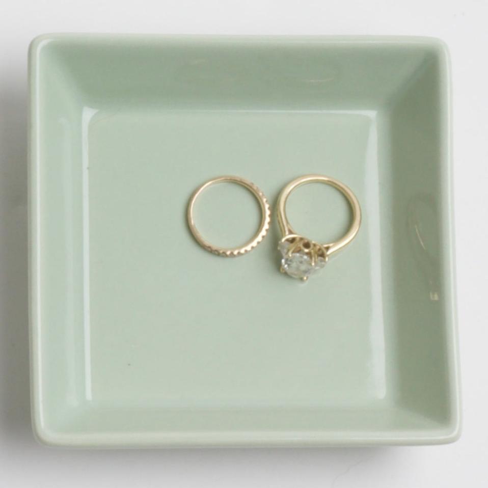 A mint-green square ceramic dish holding two gold rings. One ring is a plain gold band with a textured edge, while the other is a larger gold ring with a round, faceted gemstone, likely a diamond, in a prong setting.