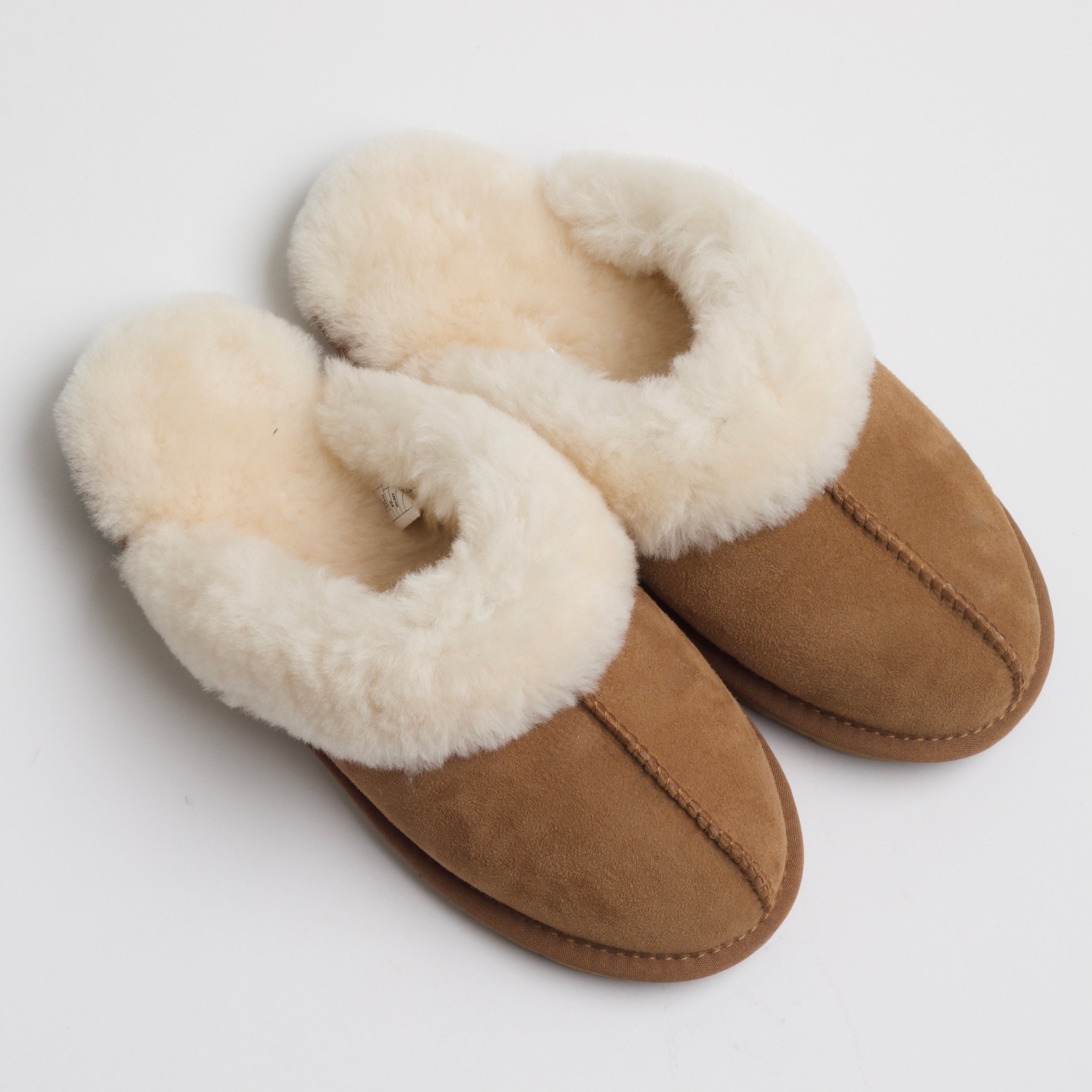 A pair of cozy, tan suede slippers lined with plush cream-colored fur. The slippers have a rounded toe and appear soft and warm, designed for indoor use. The exterior stitching is visible along the center and sides, complementing the design with a clean, natural look.