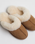 A pair of cozy, tan suede slippers lined with plush cream-colored fur. The slippers have a rounded toe and appear soft and warm, designed for indoor use. The exterior stitching is visible along the center and sides, complementing the design with a clean, natural look.