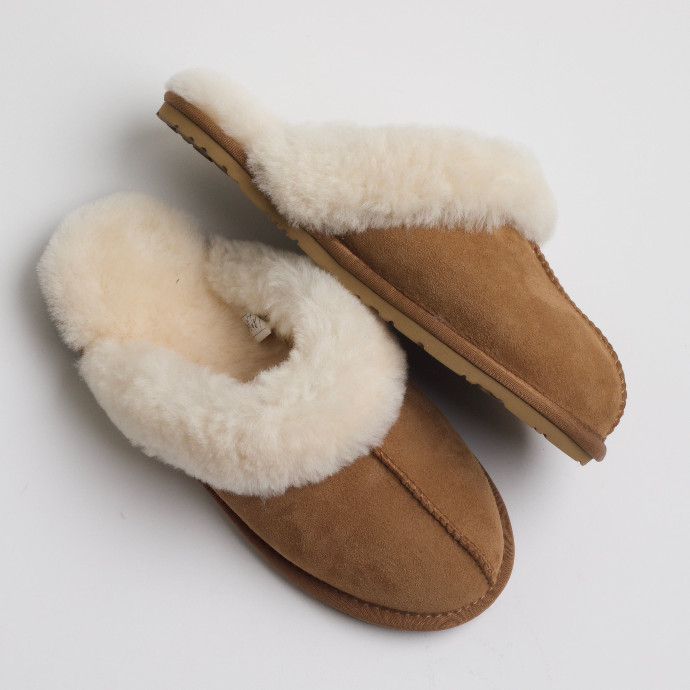 A side profile of the same tan suede slippers lined with fluffy cream fur. One slipper is laid flat, while the other is positioned on its side, showcasing the plush inner lining and the sole, which appears to be made of a durable material suitable for light indoor wear. The fur lining extends slightly over the edges, enhancing the cozy, comfortable appearance.