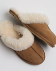 A side profile of the same tan suede slippers lined with fluffy cream fur. One slipper is laid flat, while the other is positioned on its side, showcasing the plush inner lining and the sole, which appears to be made of a durable material suitable for light indoor wear. The fur lining extends slightly over the edges, enhancing the cozy, comfortable appearance.