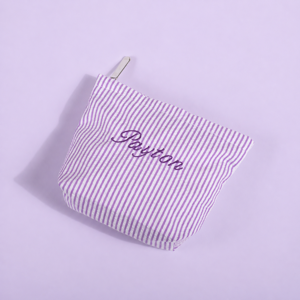A small zippered pouch with vertical lavender and white stripes, featuring the name 'Payton' embroidered in purple script on the front, set against a solid lavender background.