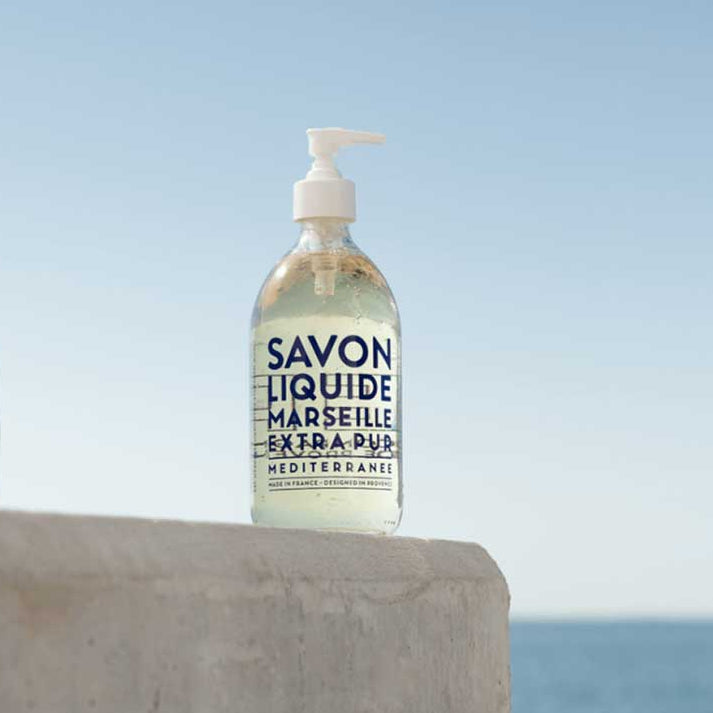 Bottle of Savon Liquide Marseille Extra Pur soap on a concrete ledge with a blue sky and ocean background.