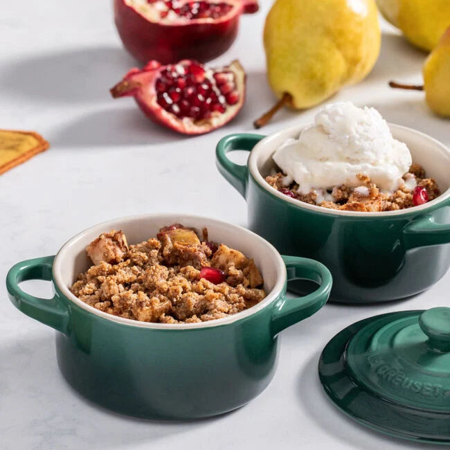 Two green ramekins with dessert on a light surface with pomegranate and pear in the background.