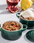 Two green ramekins with dessert on a light surface with pomegranate and pear in the background.