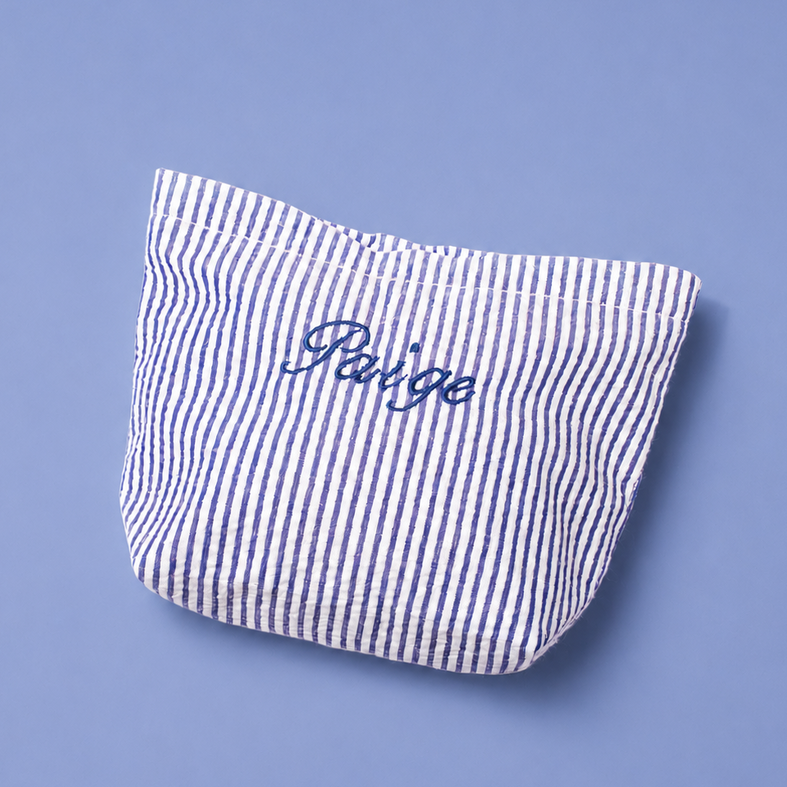 A small fabric pouch with vertical blue and white stripes and the word 'Peige' embroidered in blue script on the front, photographed on a solid periwinkle background.