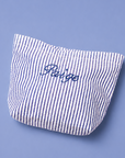A small fabric pouch with vertical blue and white stripes and the word 'Peige' embroidered in blue script on the front, photographed on a solid periwinkle background.