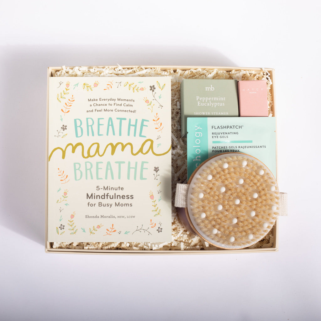 A curated gift box set for busy moms, featuring a book titled 'Breathe Mama Breathe' by Shonda Mensha, a peppermint eucalyptus diffuser, a pink bath bomb, and a natural cell resurfacer eye mask, all arranged in a kraft paper box with shredded paper filler on a white background.