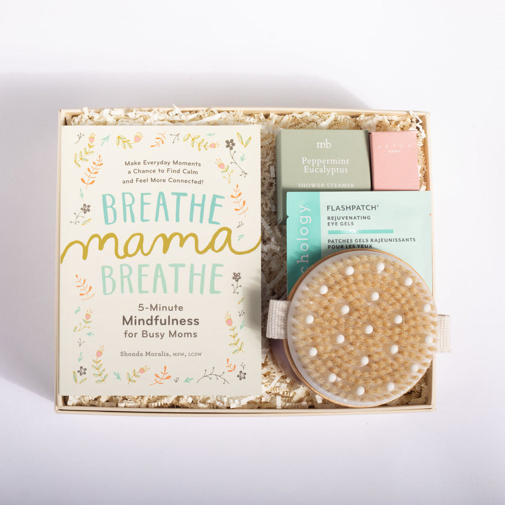 A curated gift box set for busy moms, featuring a book titled 'Breathe Mama Breathe' by Shonda Mensha, a peppermint eucalyptus diffuser, a pink bath bomb, and a natural cell resurfacer eye mask, all arranged in a kraft paper box with shredded paper filler on a white background.