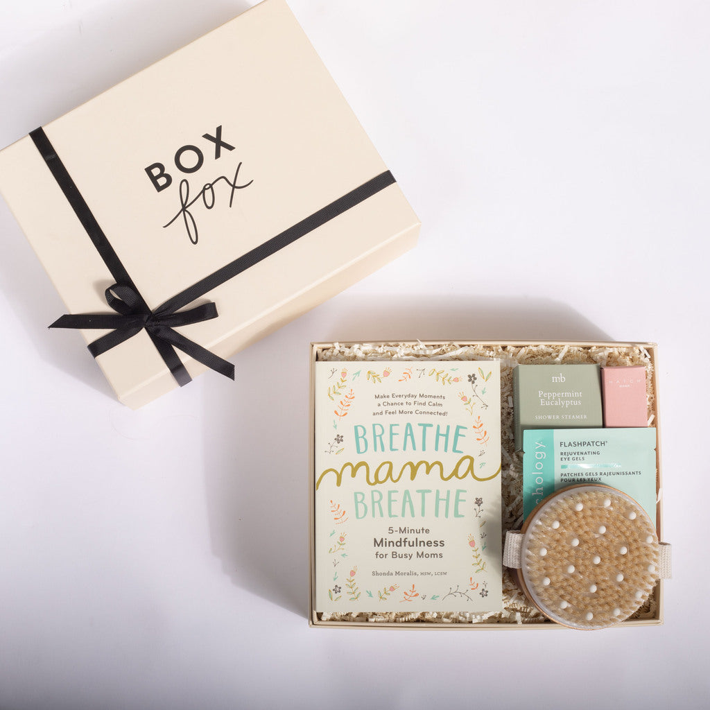 A gift set from Box Fox featuring a cream-colored box with a black ribbon, alongside an open box containing a 'Breathe Mama Breathe' mindfulness card, small bath or body items, and a round container of bath bombs, arranged on a white background.