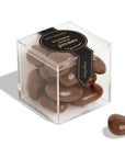 A clear plastic cube box filled with shiny chocolate-covered pecans, labeled "Off Hours Bourbon Maple Pecans" with one pecan placed outside the box on a white background.