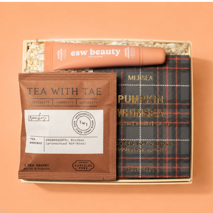 A curated gift set in a light wood box on a peach background, featuring a peach tube labeled 'esw beauty', a brown tea sachet labeled 'TEA WITH TAE', and a dark plaid pouch labeled 'MEISLA PUMPKIN WHIMSEA'.