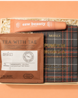 A curated gift set in a light wood box on a peach background, featuring a peach tube labeled 'esw beauty', a brown tea sachet labeled 'TEA WITH TAE', and a dark plaid pouch labeled 'MEISLA PUMPKIN WHIMSEA'.