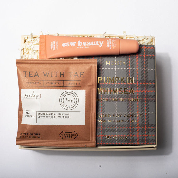 A curated gift box set containing a brown tea sachet labeled 'TEA WITH TAE', a plaid-patterned book or booklet titled 'PUMPKIN WHIMSEA', and an orange tube labeled 'esw beauty' placed on top of shredded paper inside a box, photographed against a plain light background.