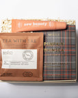 A curated gift box set containing a brown tea sachet labeled 'TEA WITH TAE', a plaid-patterned book or booklet titled 'PUMPKIN WHIMSEA', and an orange tube labeled 'esw beauty' placed on top of shredded paper inside a box, photographed against a plain light background.